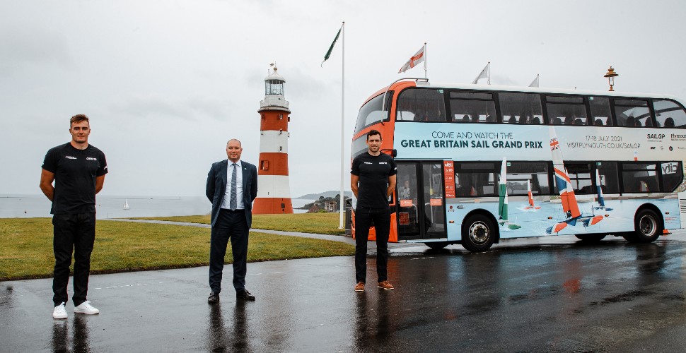 Councillor Nick Kelly and SailGP British team members Neil Hunter and Olympic rowing gold medalist Matt Gotrel on Plymouth Hoe, in front of SailGP branded bus and Smeaton's Tower 